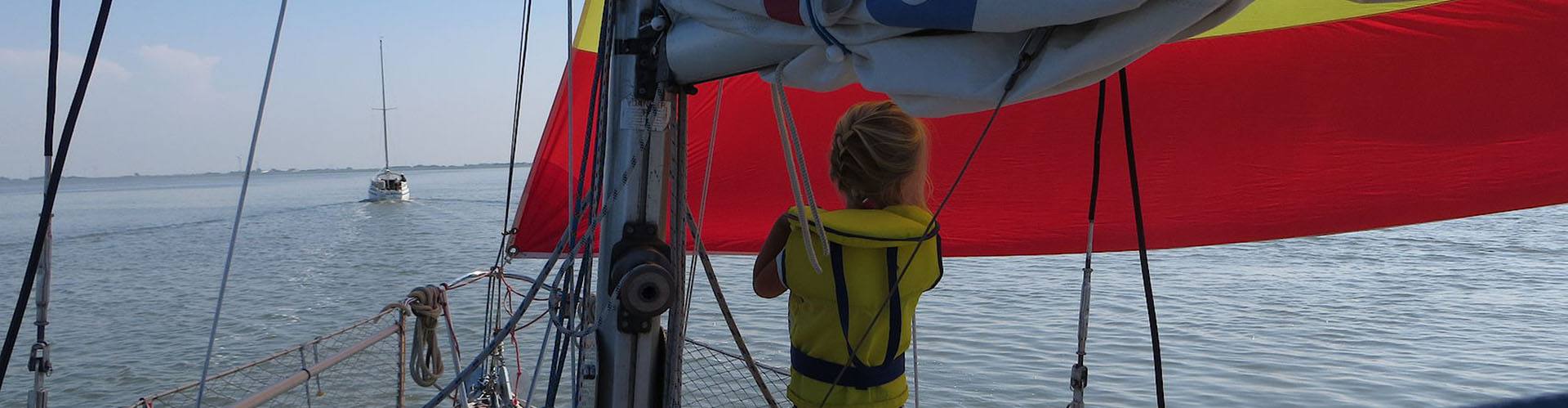 Een kind in een geel zwemvest op het voordek onder de rood-gele spinnaker, verderop vaart een andere zeilboot