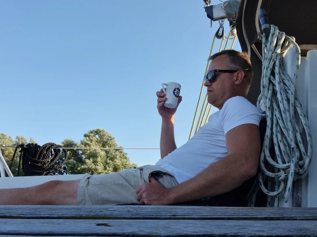 Johan ontspannen achterover op het dek, met een mok in de hand en een zonnebril op, naast opgeschoten lijnen; blauwe lucht en bomen op de achtergrond.