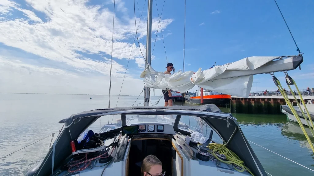 Grootzeil wordt opgedoekt in de haven van Terschelling, het wad op de achtergrond.