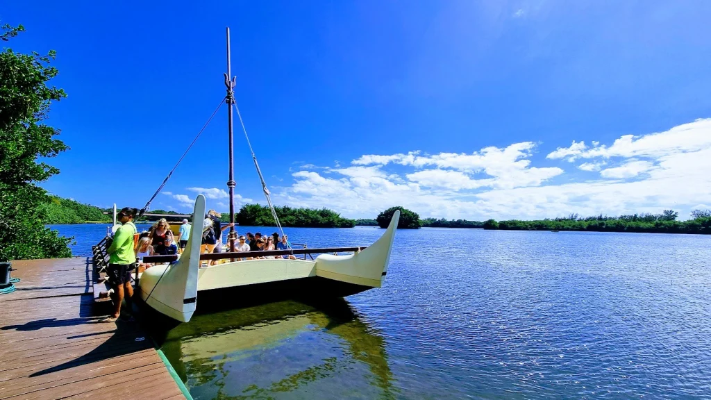 Een Polynesische dubbele kano met passagiers aan boord ligt aan een houten steiger op een blauw meer met groene oevers.