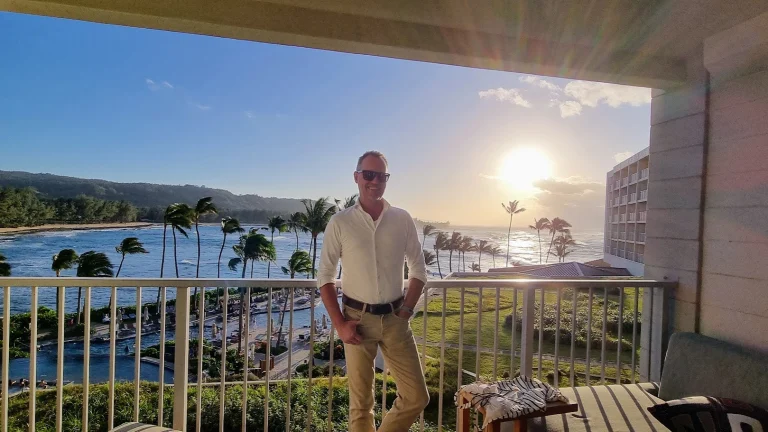 Johan op het balkon van het resort op Hawaii, met zonnebril en wit overhemd; achter hem palmbomen in de wind, zwembaden, de oceaan en de ondergaande zon.