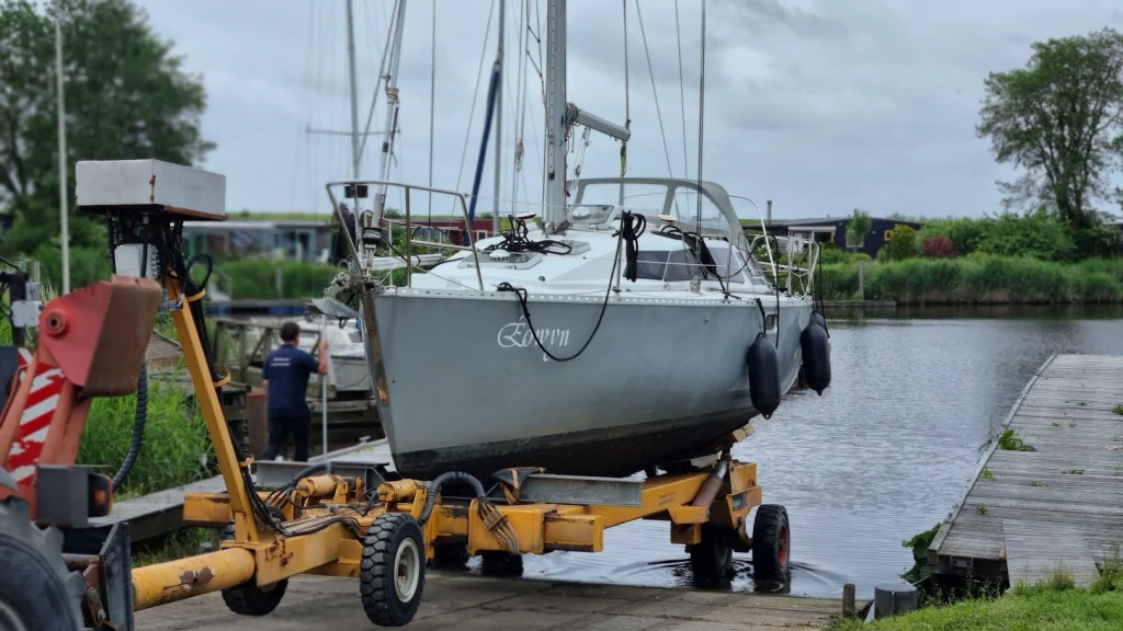 De Eowyn staat op een gele hydraulische boottrailer achter een trekker op de helling, klaar om weer te water te gaan; een werfmedewerker loopt naar de steiger, onder een grijze lucht.