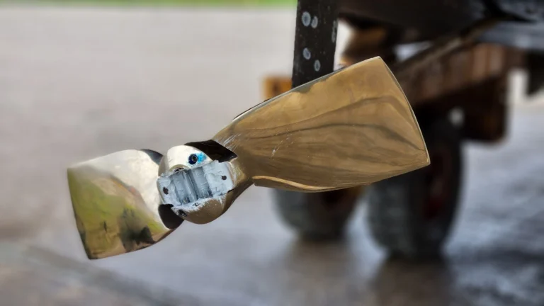 Close-up van de nieuwe, glanzend bronzen Gori-klapschroef met twee bladen, net gemonteerd onder de boot; in de naaf zit grijs vet, op de achtergrond onscherp de wielen van de boottrailer.