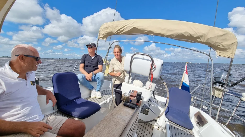 In de kuip van een Feeling 446di: moeder aan het stuurwiel, twee mannen met zonnebril op de bankjes, de Nederlandse vlag op het hek; blauwe lucht met stapelwolken boven een breed meer.