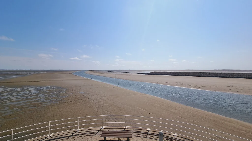 Uitzicht vanaf een uitkijkpunt met wit hek en een bankje over het drooggevallen wad, met een smalle geul met water die recht naar de horizon loopt.