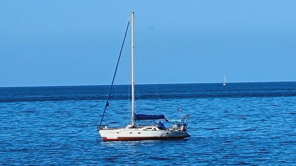 Een witte Feeling-zeilboot met rode streep ligt voor anker op diepblauw zeewater; in de verte een tweede zeilboot aan de horizon.