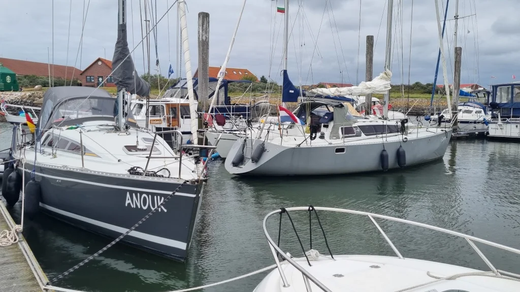 In de jachthaven van Baltrum: een grijze Feeling 346 tussen andere jachten, vooraan de donkergrijze zeilboot Anouk; bewolkte lucht en rode daken op de dijk.