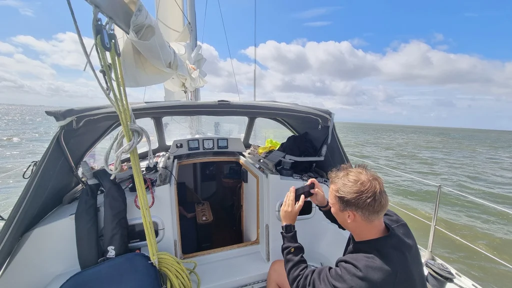 Mijn neefje fotografeert met zijn telefoon de instrumenten bij de kajuitingang; zon, stapelwolken en ruim water rondom.