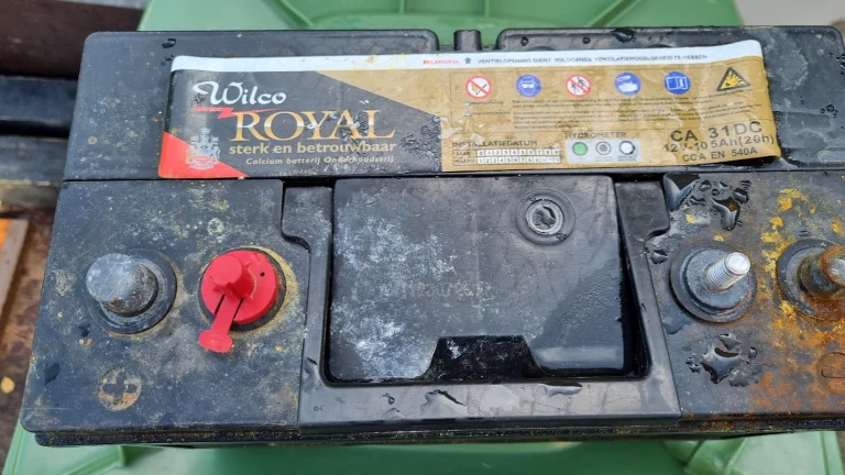 Een van de oude accu's uit de boot, van bovenaf: een vuile Wilco Royal calciumaccu van 12 V 105 Ah met een rode poolkap, waterdruppels en roestbruine aanslag rond de polen.
