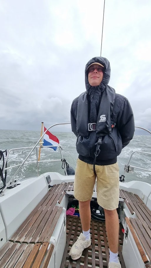 Mijn neefje staat achter in de kuip in een zwarte jas met capuchon op en een reddingsvest om, glimlachend in de wind; grijze lucht en woelig water achter hem.
