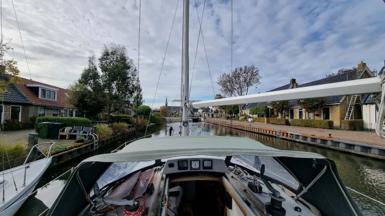 Vanuit de kuip van de Eowyn, onder de groene buiskap door, over het voordek: de boot vaart met staande mast door een smalle dorpsvaart tussen oude huisjes, richting een ophaalbrug met daarachter een kerktoren, onder een lucht vol schapenwolkjes.