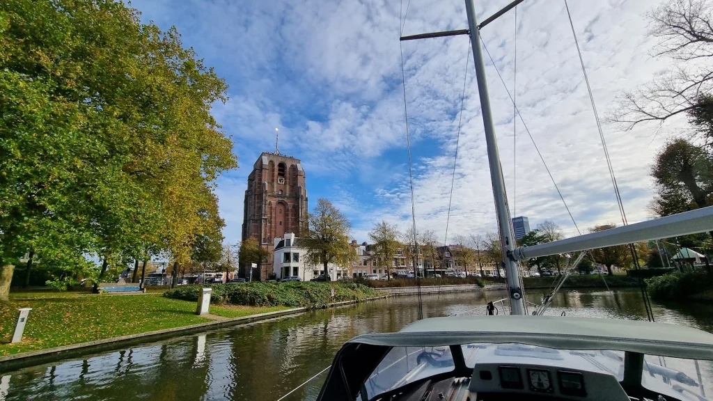 Door Leeuwarden: vanaf de boot zicht op de scheve Oldehove-toren boven de bomen in herfstkleuren, aan het water van de stadsgracht, onder een blauwe lucht met wolken.