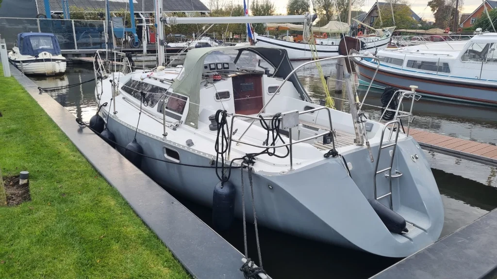 De Eowyn, een grijze zeilboot met groene buiskap, ligt met stootwillen netjes afgemeerd in een box langs een kade met gras; op de achtergrond een blauwe bootlift en een traditioneel zeilschip.