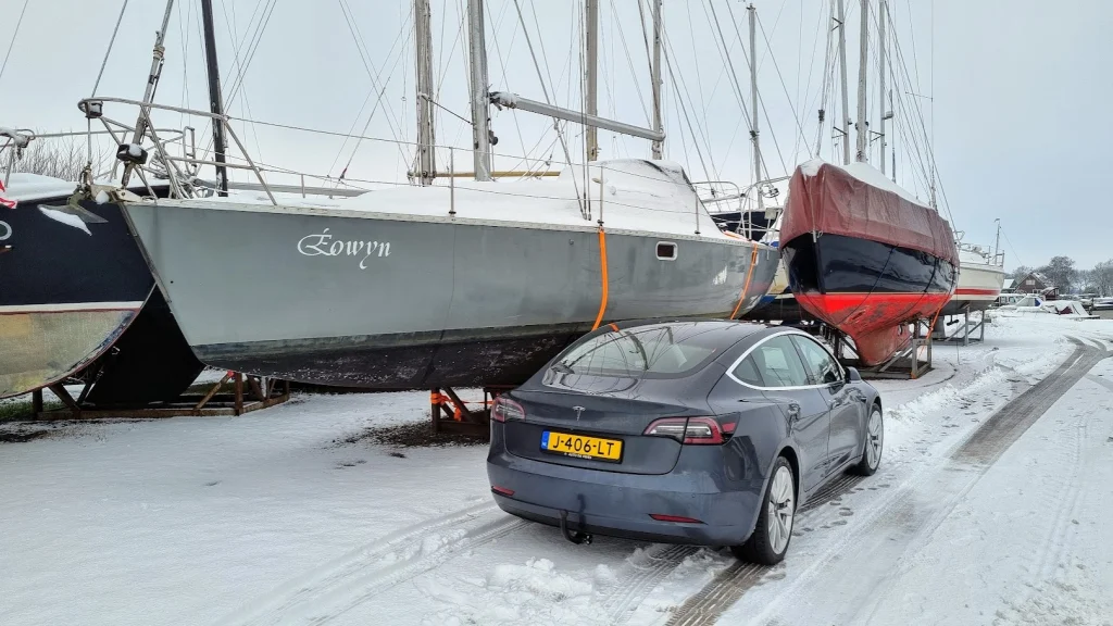 De Eowyn staat in de winterstalling op de wal, op een bok tussen andere zeilboten, met sneeuw op het dek en op het terrein; ervoor staat mijn grijze Tesla in de sneeuw.
