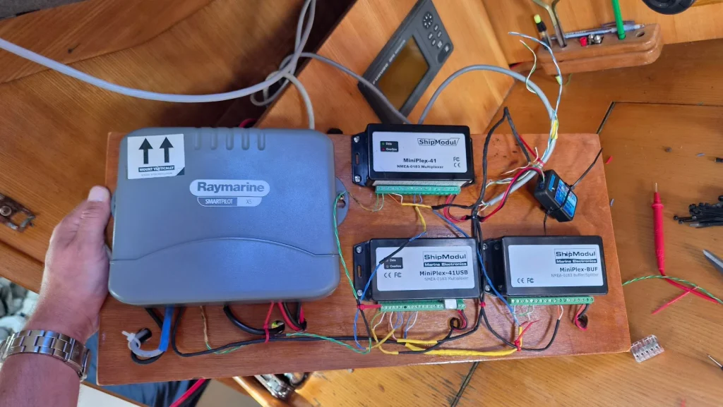 Een hand houdt een houten plank vast waarop de scheepselektronica is gemonteerd en bedraad: een grijze Raymarine SmartPilot X5-stuurautomaatcomputer en drie zwarte ShipModul-kastjes (MiniPlex-41, MiniPlex-41USB en MiniPlex-BUF) voor NMEA-0183-data, met een wirwar van gekleurde draadjes; op de achtergrond de kaarttafel met een GPS-scherm en gereedschap.
