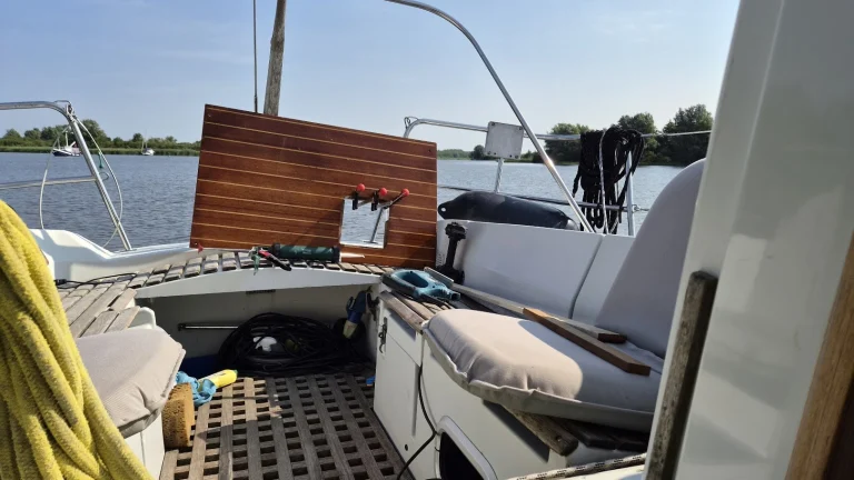 De kuip van de Eowyn voor anker op een rustig meer in de zon: een houten paneel met lichte biezen, met een uitgezaagd gat en drie lijmklemmen, staat rechtop tegen de achterbank; ernaast een kitpistool en een decoupeerzaag, in de kuip een verlengkabel en een spons.