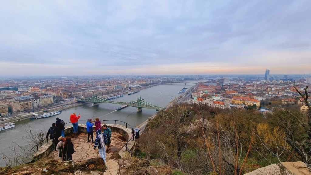 Boedapest: uitzicht vanaf een uitkijkpunt op de Gellértheuvel over de Donau, met de groene Vrijheidsbrug, rondvaartboten en de daken van de stad tot aan de horizon; op het stenen plateau op de voorgrond staan wat toeristen.
