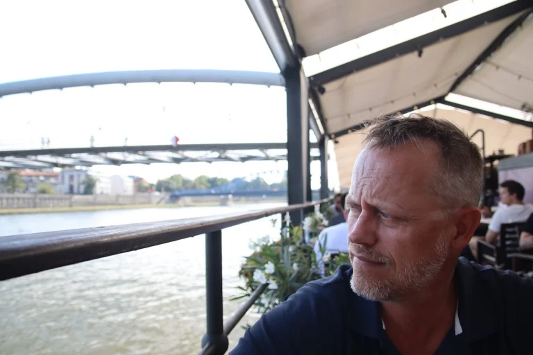 Krakau: een man met grijzend kort haar en baardje kijkt bedenkelijk opzij vanaf een overdekt terras aan de Wisła; achter hem over het water de boogbrug voor voetgangers en in de verte een blauwe spoorbrug.