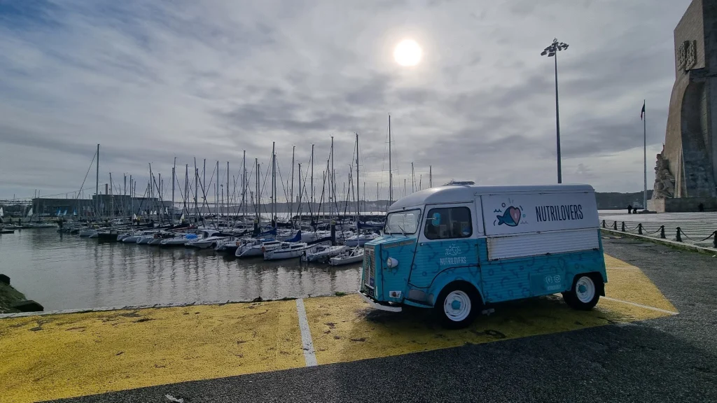 Lissabon: een lichtblauw-wit Citroën-busje als foodtruck staat aan de jachthaven van Belém met tientallen zeilboten, naast het grote stenen Monument voor de Ontdekkingen, met een waterig zonnetje achter de wolken.