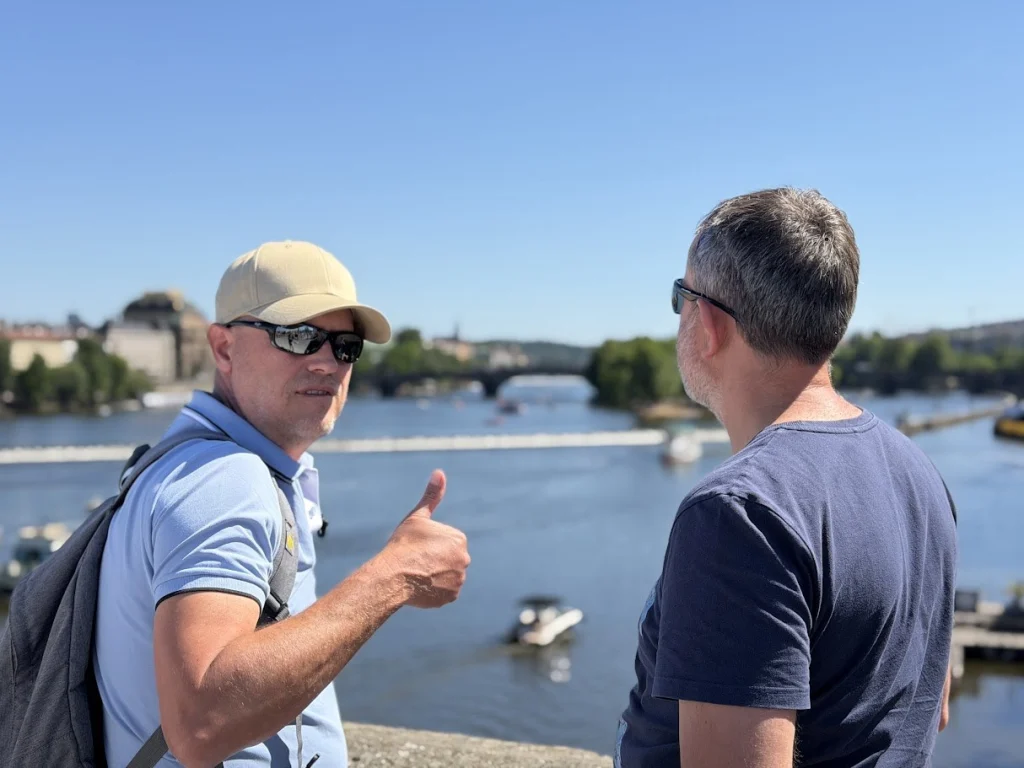 Praag: twee mannen met zonnebril staan aan de Moldau; de een, met pet en rugzak, steekt zijn duim op naar de camera. Op de rivier een stuw, bootjes en in de verte een stenen brug, onder een strakblauwe lucht.