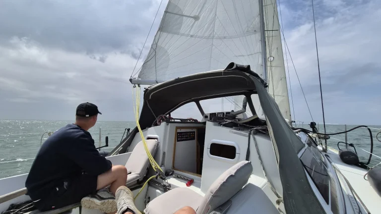 Aan de wind op het wad: een bemanningslid met zwarte pet en donkere trui zit op de kuiprand en kijkt over het glinsterende water; de fok en het grootzeil staan bol, de buiskap is half open.