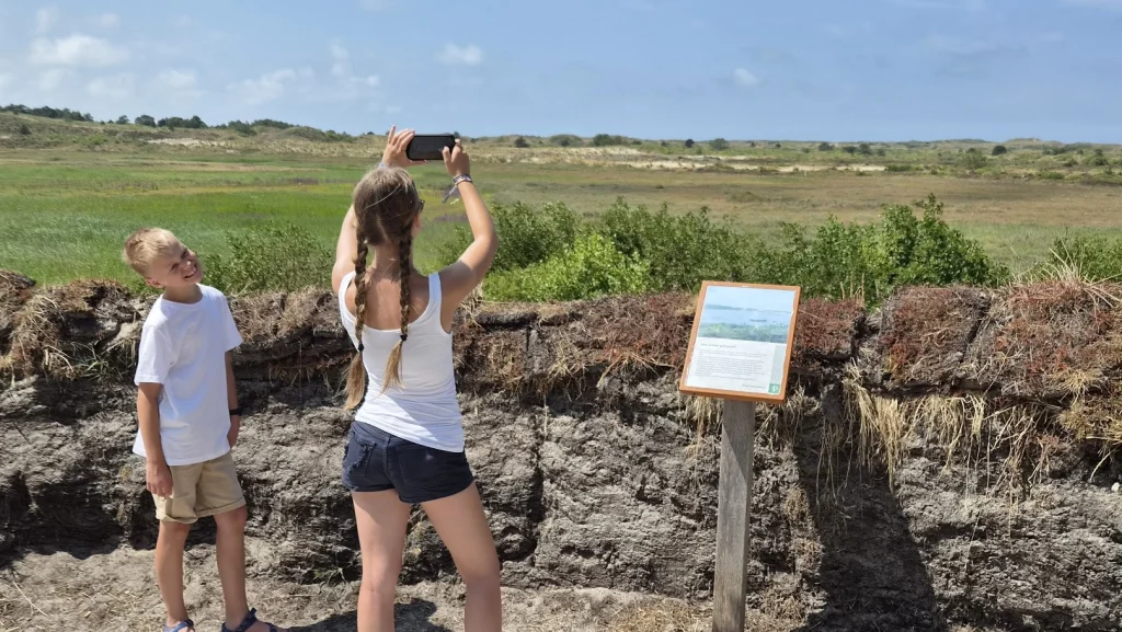 De kinderen bij een wand van opgestapelde plaggen met een informatiebord in de duinen: het meisje met lange vlechten maakt met haar telefoon een foto van de groene vallei met duinen, de jongen staat ernaast en lacht.