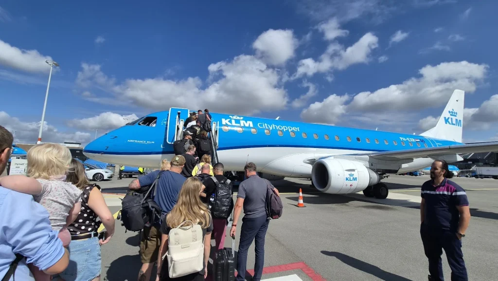 Passagiers met rugzakken staan in de rij op het platform om via de trap een blauw-wit KLM Cityhopper-vliegtuig in te stappen, onder een blauwe lucht met witte wolken.
