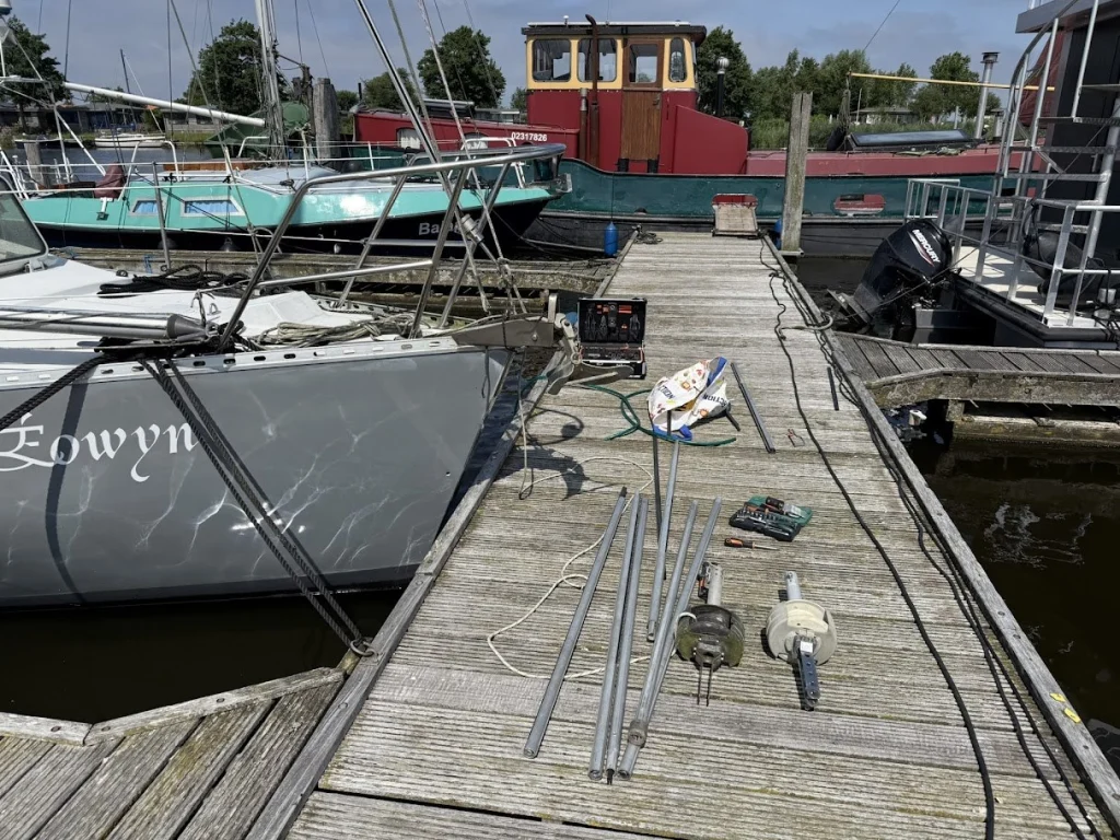 Op de steiger naast de boeg van de Eowyn liggen de onderdelen van de rolfok uitgestald: lange aluminium profielstukken, twee rolfoktrommels, een gereedschapskoffer en schroevendraaiers.