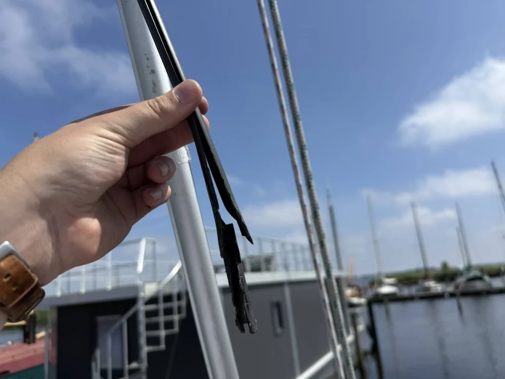 Een hand houdt het kapotte zwarte kunststof binnenprofiel van de rolfok omhoog, gerafeld en afgebroken, naast het aluminium voorstagprofiel, met de haven op de achtergrond.