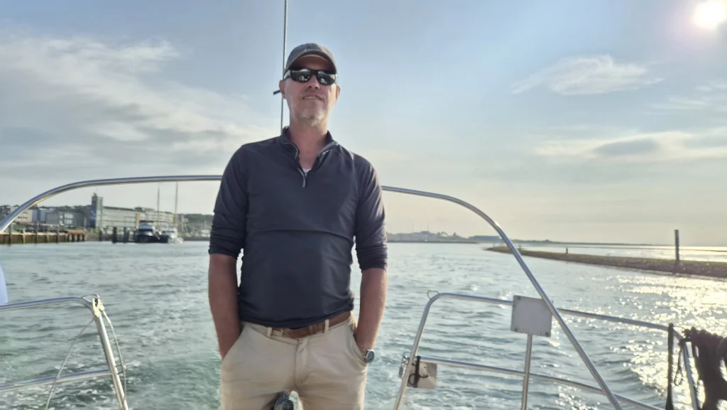 Een man met pet en zonnebril staat met zijn handen in de zakken op het achterdek tussen de hekstoelen, terwijl de boot een eilandhaven uitvaart; achter hem de haven en een strekdam in het avondlicht.