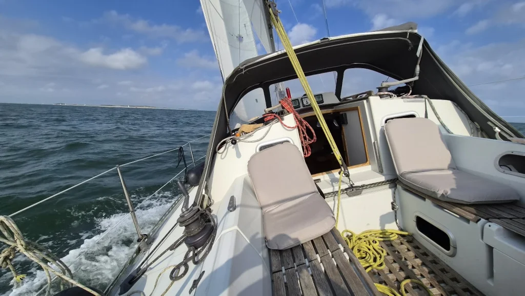Zeilen op de Waddenzee: vanuit de kuip met de lege kussens zie je de boot flink hellen, het water schuimt langs de romp en in de verte ligt een eiland onder een blauwe lucht met stapelwolken.